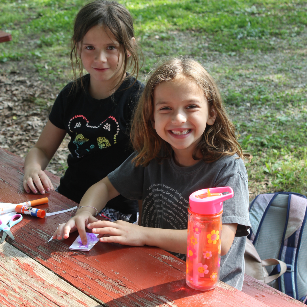 Photo from J Day Camps 2025 at The J - St. Louis: Two girls of about 10 sit at a picnic table, drinking water and crafting.