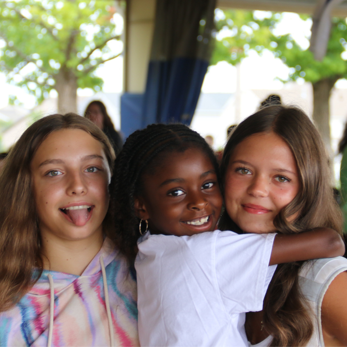 Photo from J Day Camps 2025 at The J - St. Louis: 3 campers, girls of about 10 or 11, hug each other, smiling at the camera