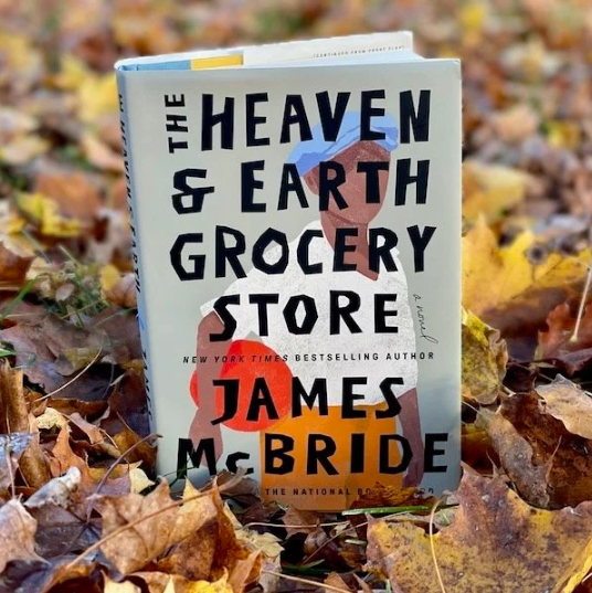 A hardcover book, The Heaven and Earth Grocery Story, by James McBride, stands on the ground among a layer of fallen autumn leaves.